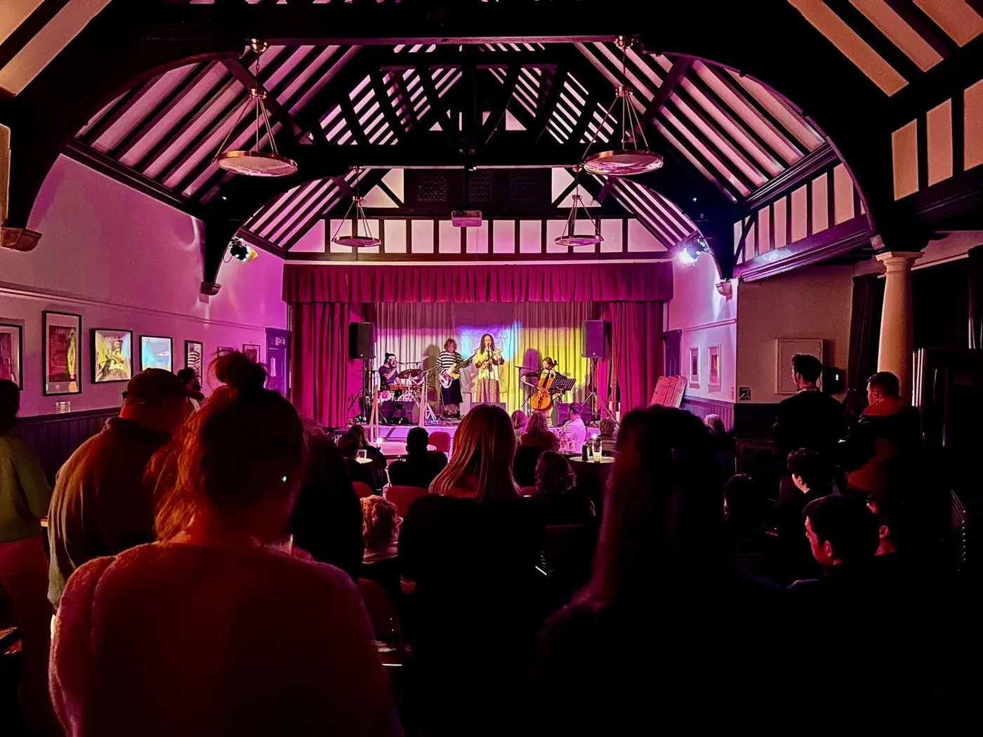 A band with a cellist playing on a lit stage under a timbered roof, watched by a seated and standing audience