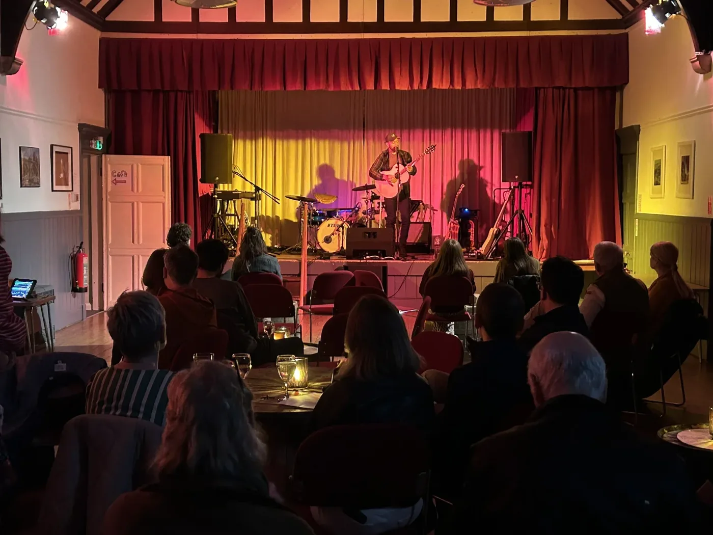 A solo guitarist on a stage with red and gold curtains, playing to an audience seated at small candlelit tables